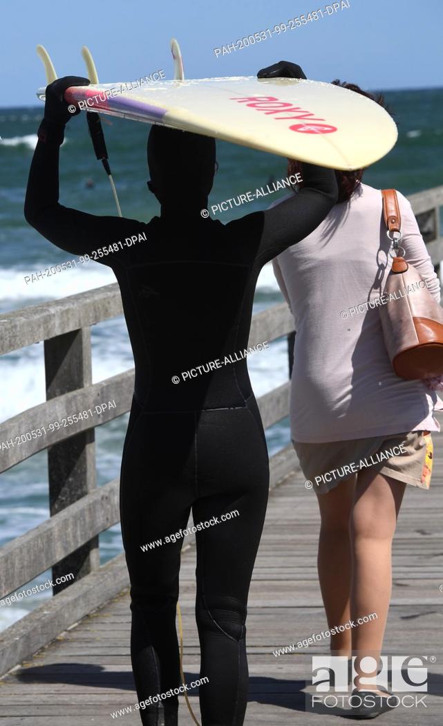 Stock Photo: 31 May 2020, Mecklenburg-Western Pomerania, Binz: A woman is carrying a surfboard on the pier next to passers-by walking across the pier.