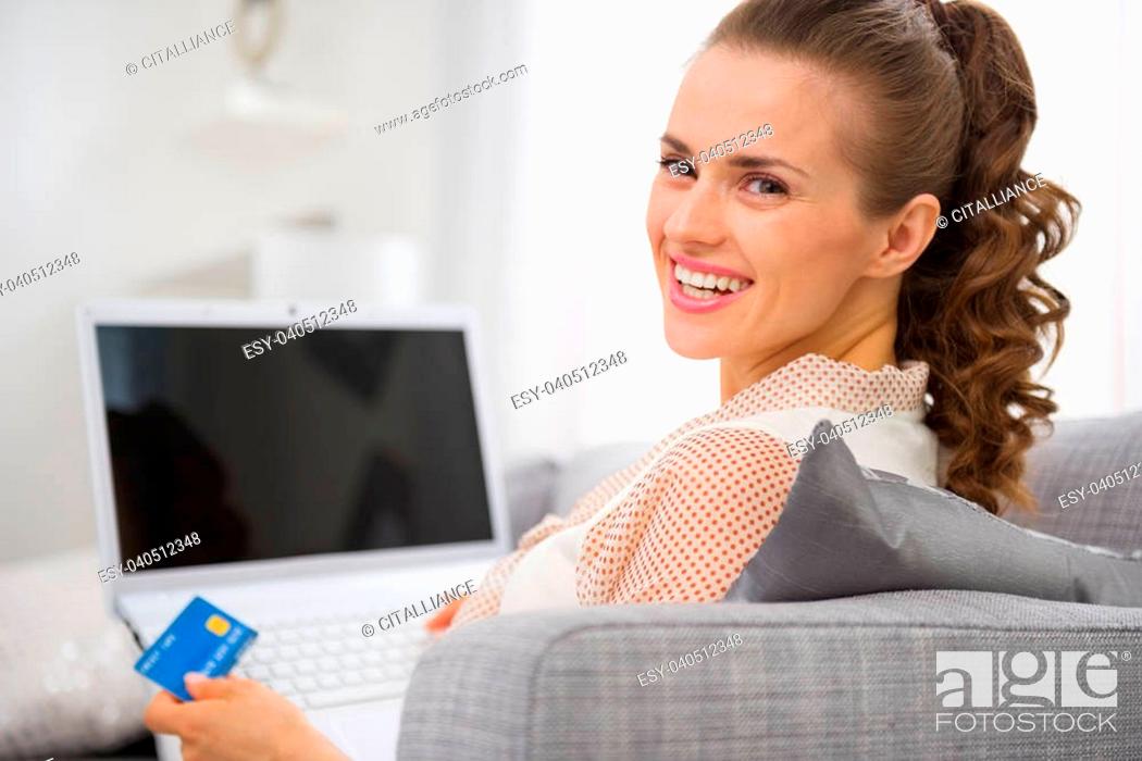 Stock Photo: Smiling young housewife with laptop and credit card sitting on couch.