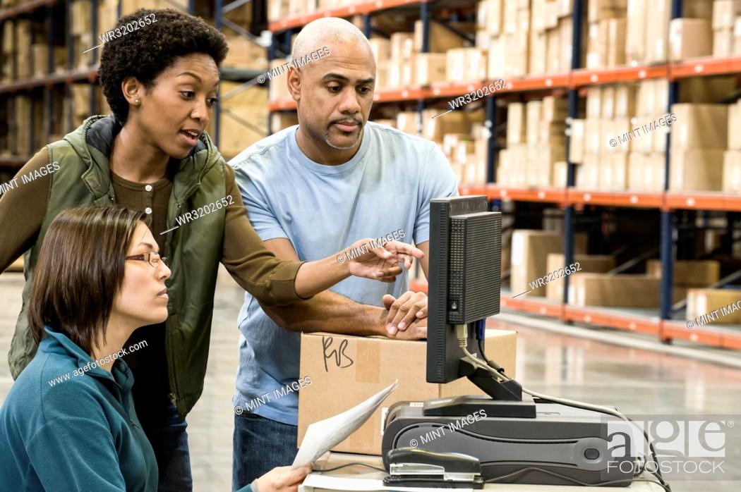 Stock Photo: A group of three mixed race warehouse workers checking inventory on a computer in a large distribution warehouse.