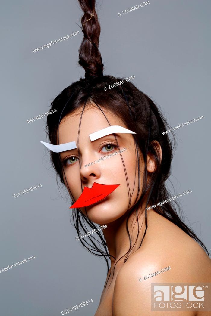 Stock Photo: Beautiful young woman with odd fancy hairstyle and eyebrows and lips paper cutouts on face. Beauty shot on grey background. Copy space.