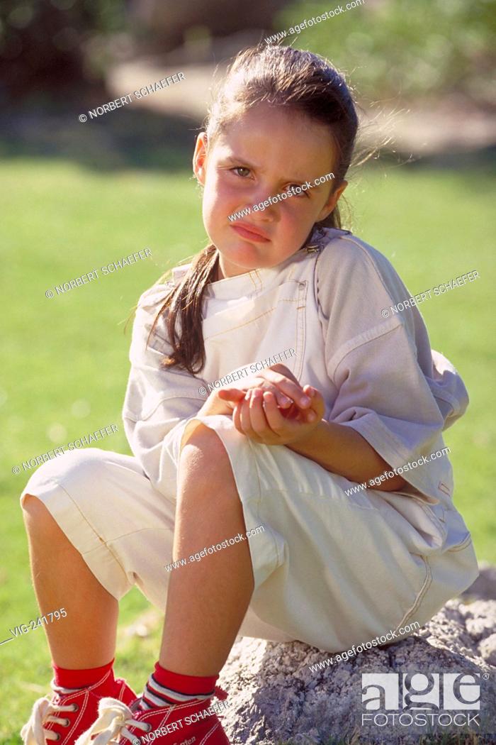 Stock Photo: portrait, outdoor, sad looking 8-year-old girl with long brown hair wearing white short trousers and red basketball shoes sits on a stone in a park - GERMANY.