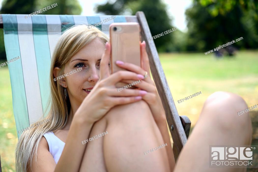 Stock Photo: Young blonde woman in London Hyde Park, sitting in a beach chair and enjoying the late summer sun and browsing the internet on her mobile phone.