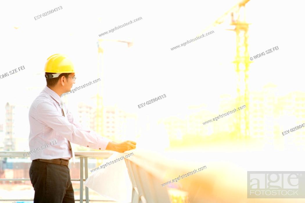 Stock Photo: Asian Indian male site contractor engineer with hard hat holding blue print paper inspecting at construction site, crane with golden sunlight at the background.