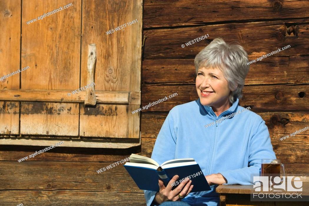 Stock Photo: Austria, Senior woman reading book by log cabin, portrait.