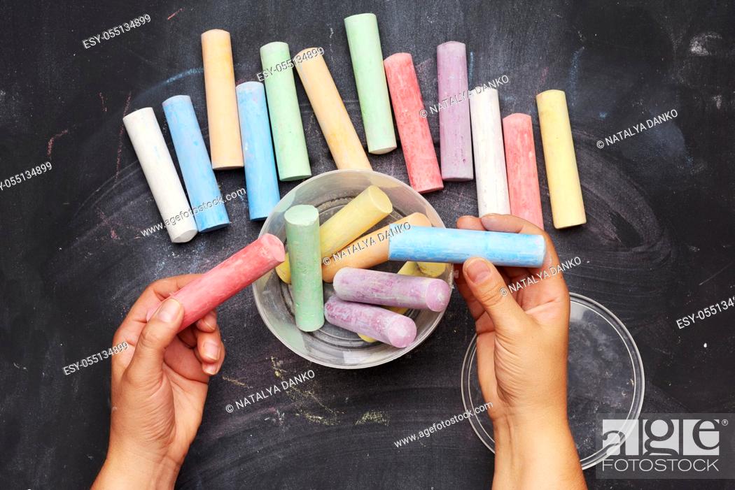 Imagen: multicolored chalk in a plastic bucket on a black background, female hands fold objects into a container, top view.