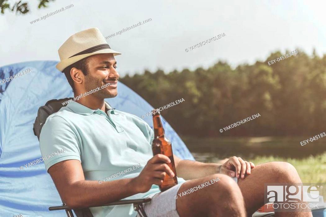 Stock Photo: Delighted guy enjoying the nature while sitting on folding chair and holding bottle of beer. Forest and tent on background.