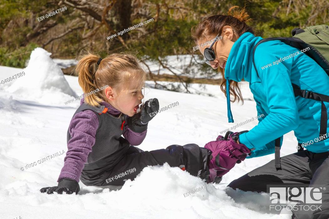 Stock Photo: woman placing boot to her funny daughter, four years old blonde girl, sitting on the snow in nature. Child is eating snow with her hand.