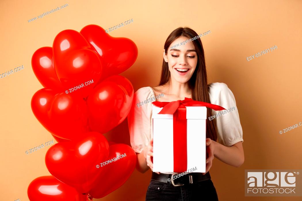 Stock Photo: Happy woman open Valentines day gift, smiling excited and looking at present box, standing near red heart balloons on beige background.