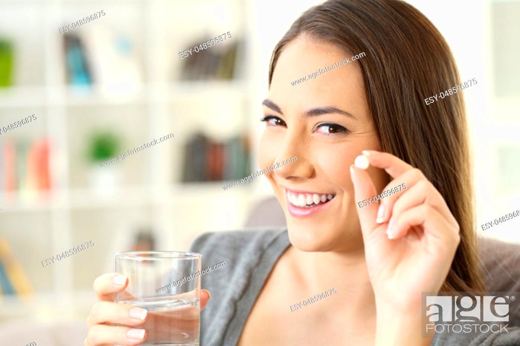 Stock Photo: Happy woman holding a white round pill and a glass of water looking at camera sitting on a sofa in a house interior.