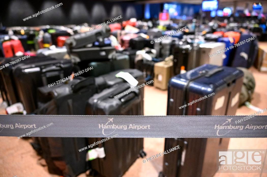 Stock Photo: 23 June 2022, Hamburg: ""Hamburg Airport"" is written on a barrier tape in front of numerous stored suitcases in the baggage claim area.