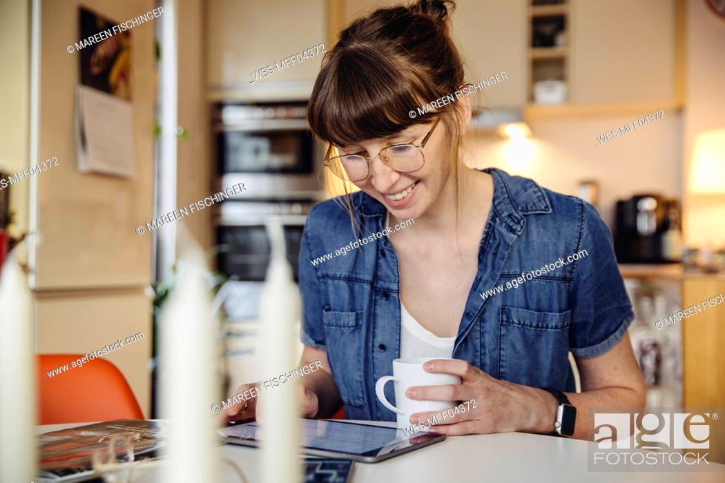 Stock Photo: Smiling woman sitting with cup of coffee at table in the kitchen using tablet.