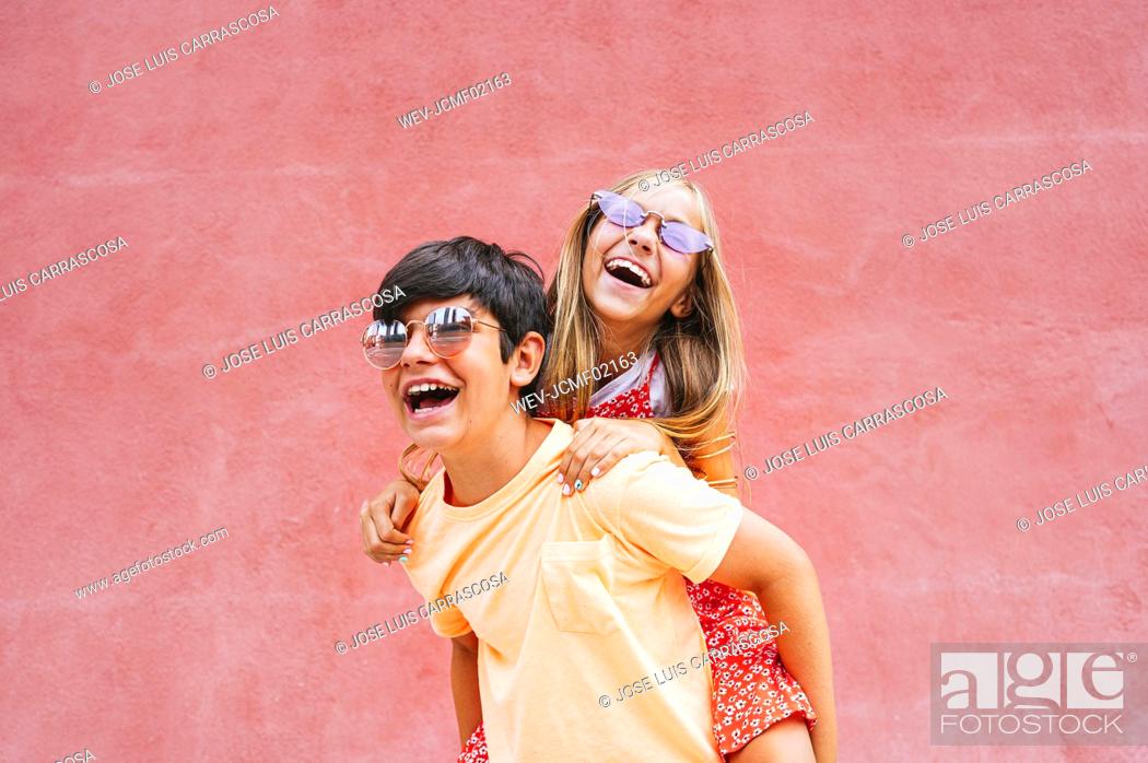 Stock Photo: Cheerful boy piggybacking female in front of red wall.