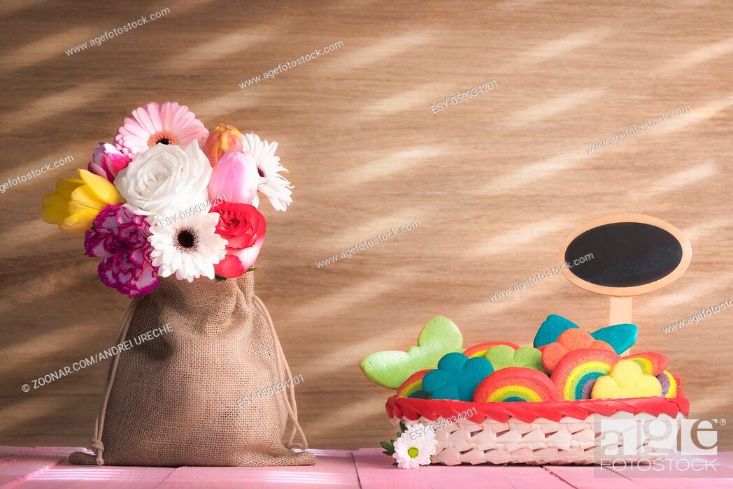Stock Photo: Bouquet of diverse flowers in a jute sack, a basket full of colorful and multi-shaped cookies, with a blank banner, on a pink table, on a sunny day.
