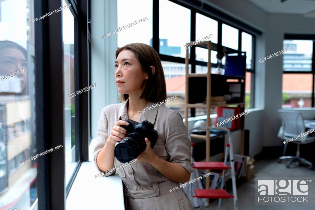 Stock Photo: Asian businesswoman holding camera looking through window in creative office.