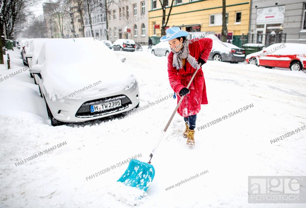 Stock Photo: 08 February 2021, Lower Saxony, Hanover: Lek, dressed in a red bathrobe and blue hat, frees a driveway on Sall Street in South Town.