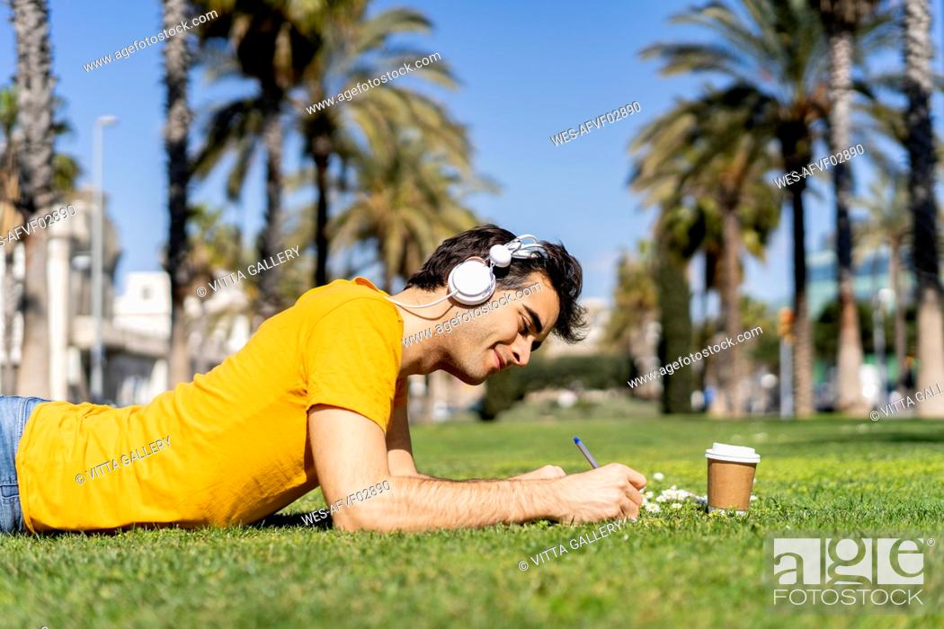 Stock Photo: Spain, Barcelona, man lying on lawn in the city with headphones and notebook.