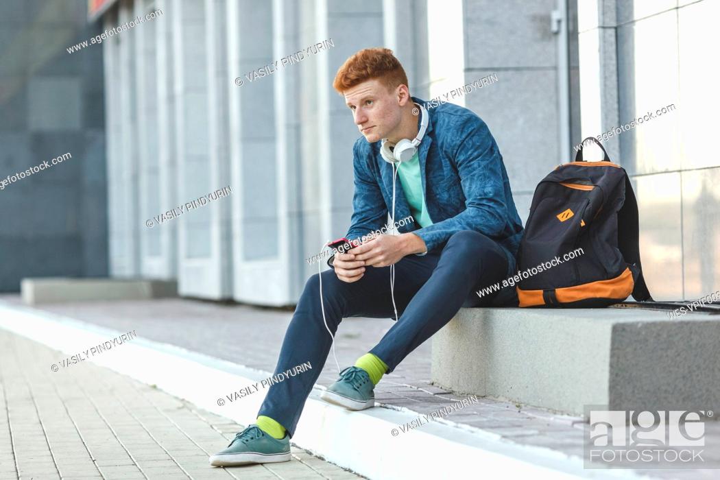 Stock Photo: Redheaded young man sitting outdoors with smartphone and headphones.