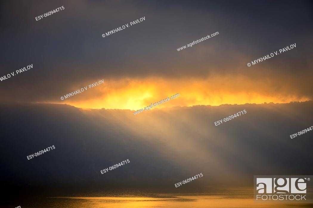 Stock Photo: Heavy clouds over the rocky shore. Sunrays barely break through, illuminate small houses and the water surface.