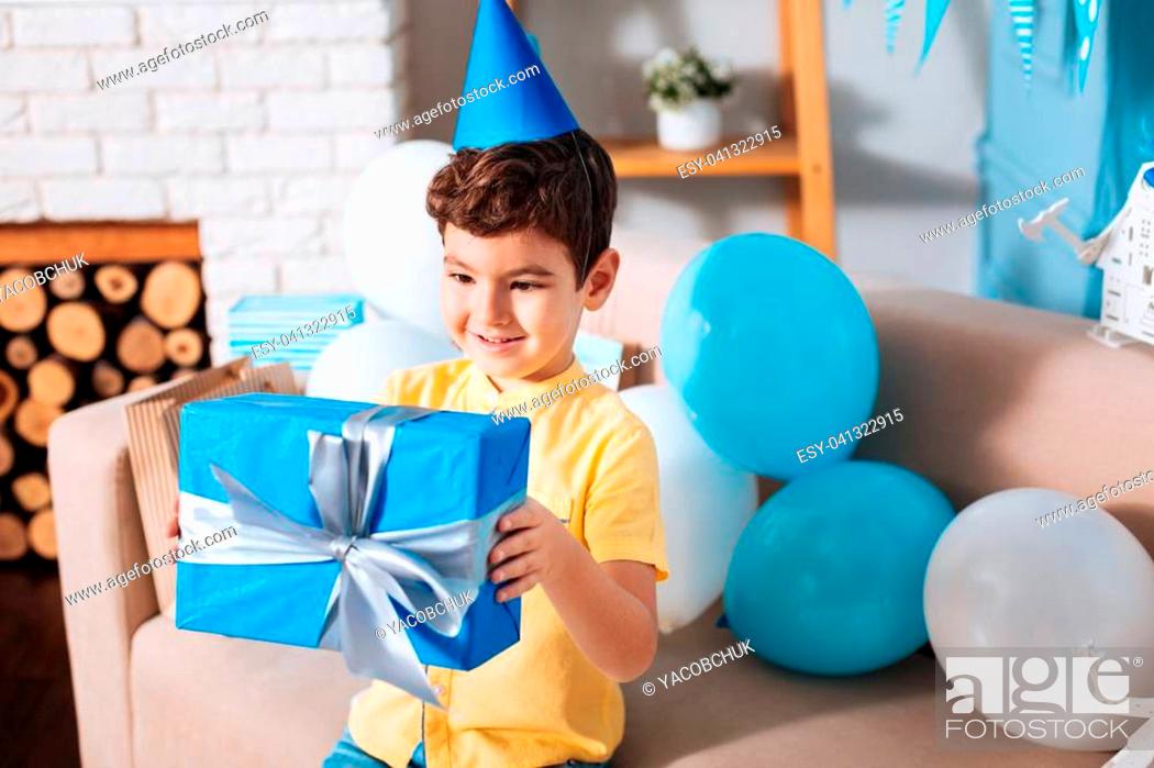 Stock Photo: Childlike curiosity. Cute little boy examining a blue box with his birthday present and smiling while sitting on the sofa full of balloons.