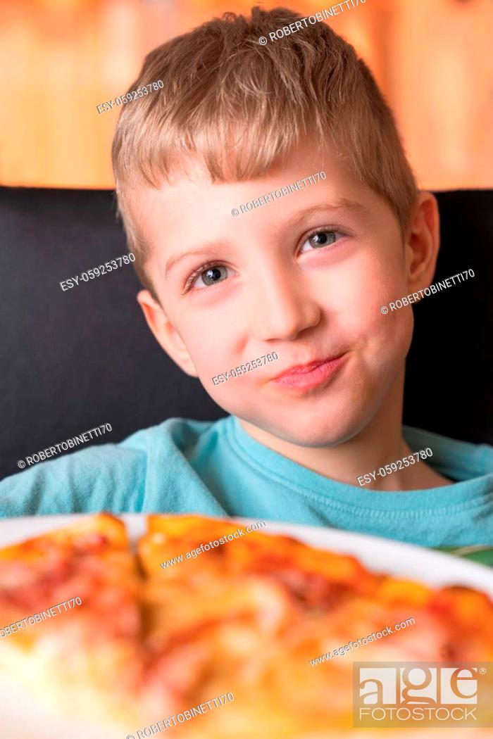 Stock Photo: Beautiful happy young boy smiling and eating fresh made pizza, He sit at black chair, He has blonde hair.