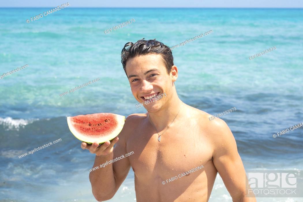 Photo de stock: Young fit body boy portrait holding red fresh watermelon with blue ocean water in background. Summer holiday vacation season concept lifestyle people.