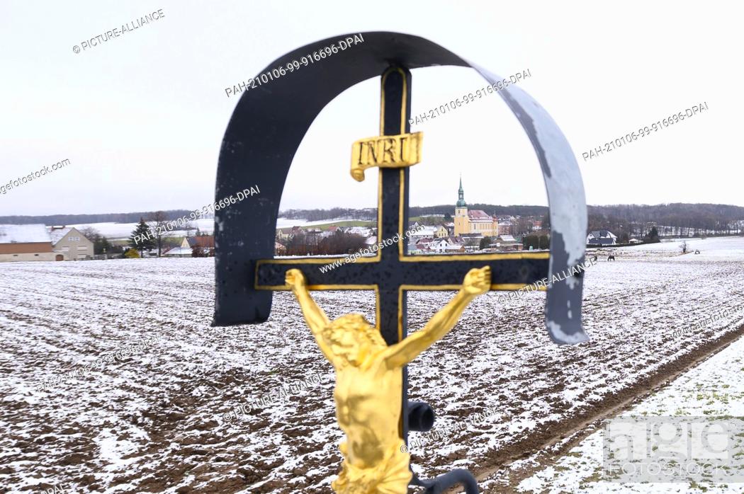 Photo de stock: 06 January 2021, Saxony, Crostwitz: A cross stands in front of a snow-covered field at the entrance to the village; the parish church can be seen in the.