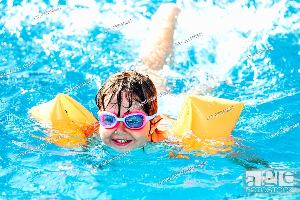Stock Photo: Little black-haired girl smiling and swimming in the pool wearing sleeves and swimming goggles.