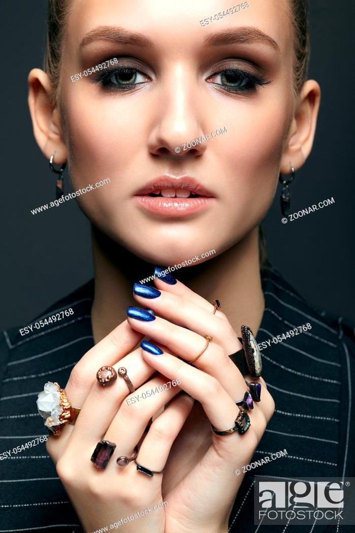 Stock Photo: Beauty portrait of young female with blue manicure on dark background. Young beautiful woman with many bijouterie rings with stones on fingers.