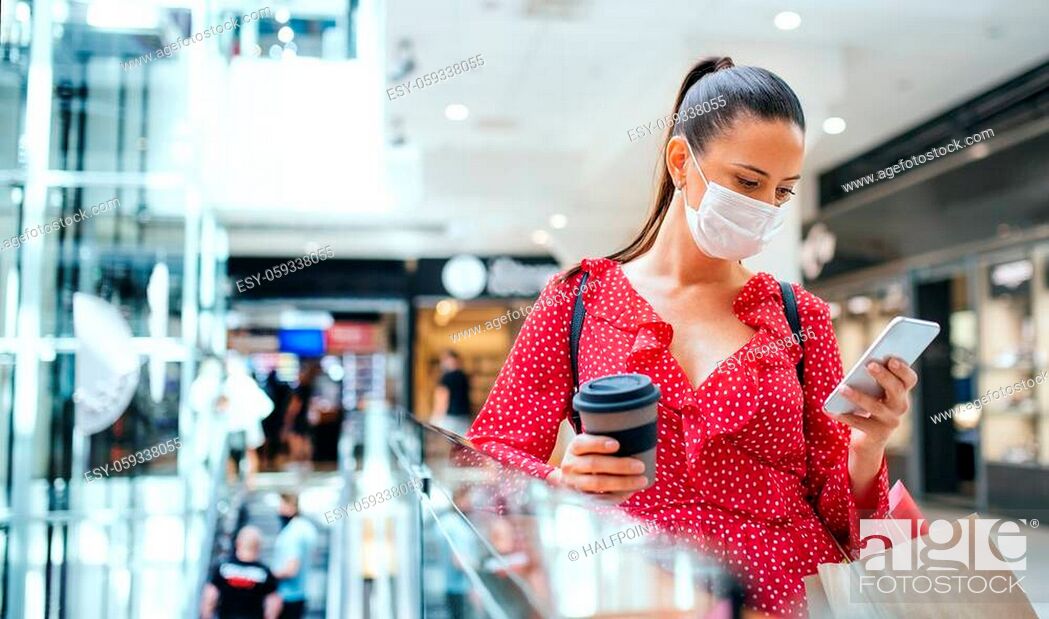 Stock Photo: Young woman with face mask standing and using smartphone indoors in shopping center, coronavirus concept.