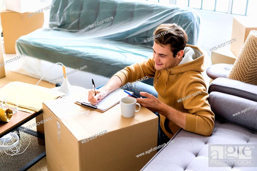 Stock Photo: Smiling man with mobile phone writing on paper over cardboard box in new home.