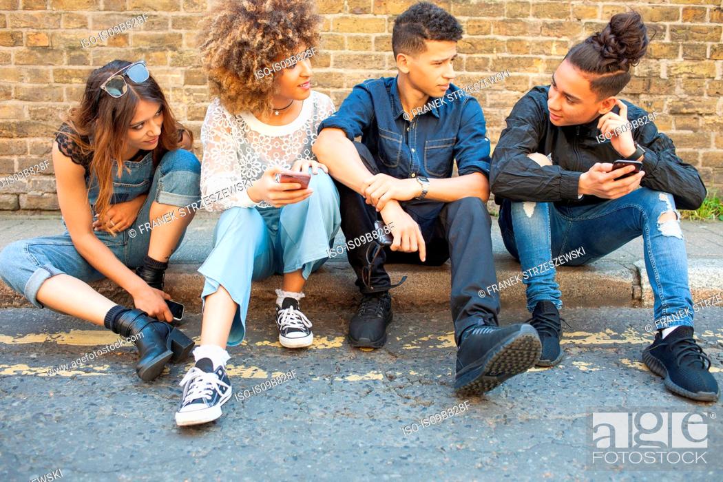 Stock Photo: Four friends sitting in street, looking at smartphones.