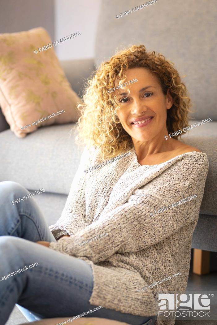 Stock Photo: Portrait of happy young woman sitting on floor at home. Beautiful caucasian woman smiling sitting in living room. Pretty young woman with curly hair sitting in.