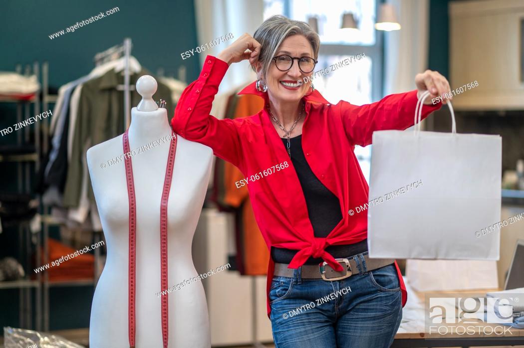 Stock Photo: Determination. Gray-haired elegant woman smiling showing package at camera standing leaning on mannequin in atelier.