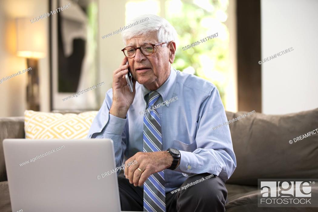Stock Photo: Mature Caucasian Male doctor practicing tele-medicine from his home, using cell phone and laptop computer, Talking to patient.