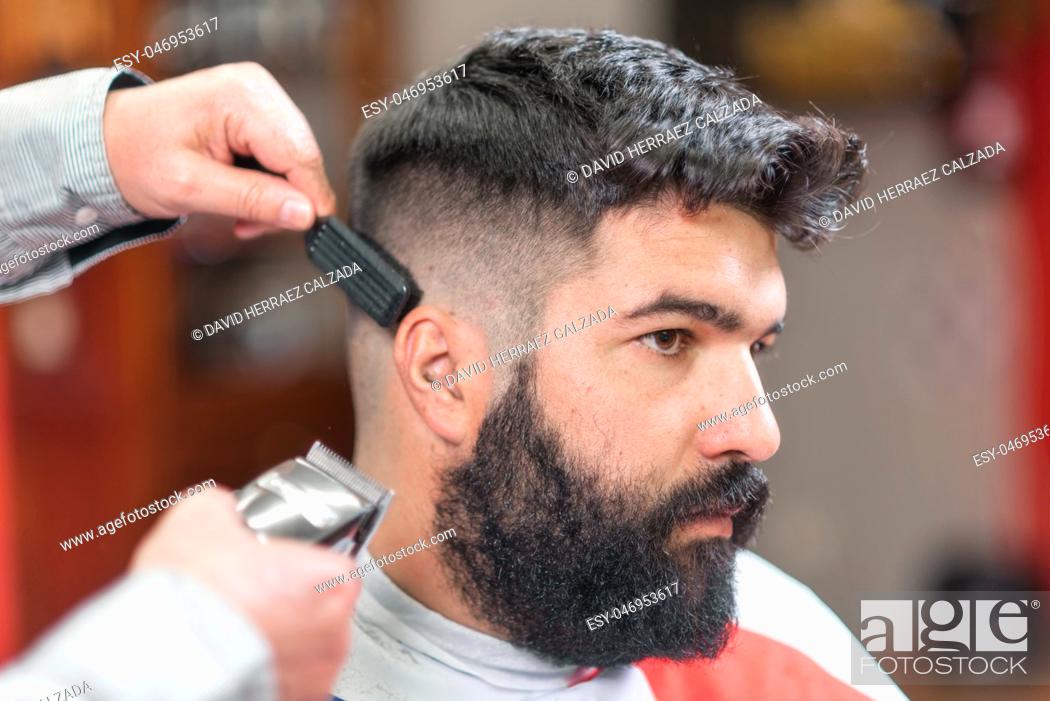 Stock Photo: Handsome bearded man, getting haircut by barber, with electric trimmer at barbershop .