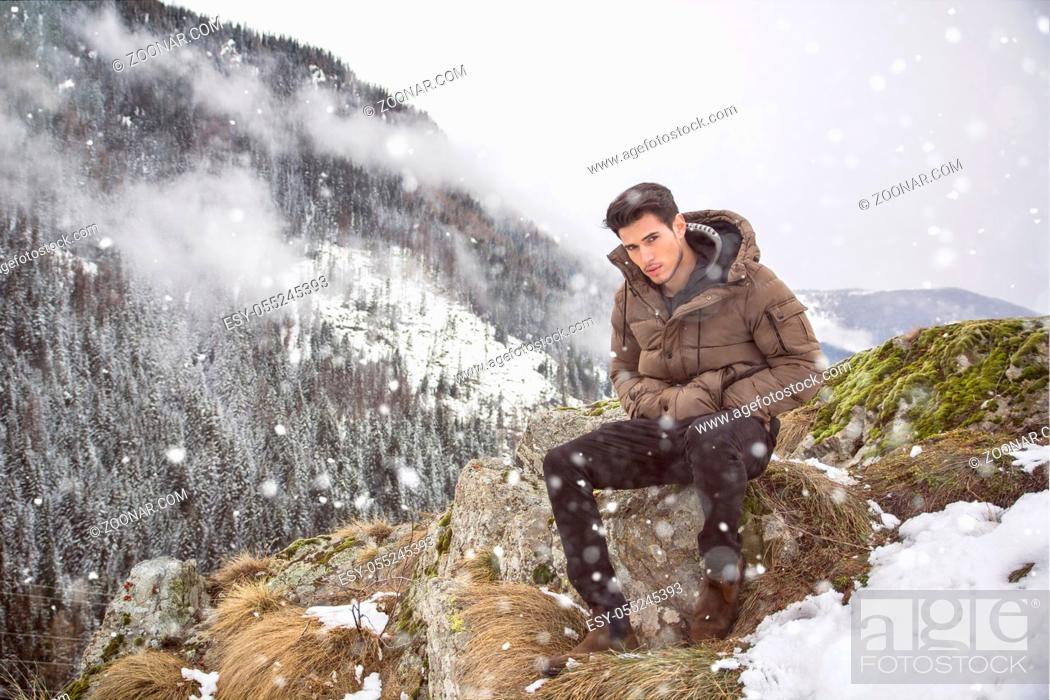 Stock Photo: Handsome young man in winter outerwear sitting under snow up the mountain, while looking at camera.