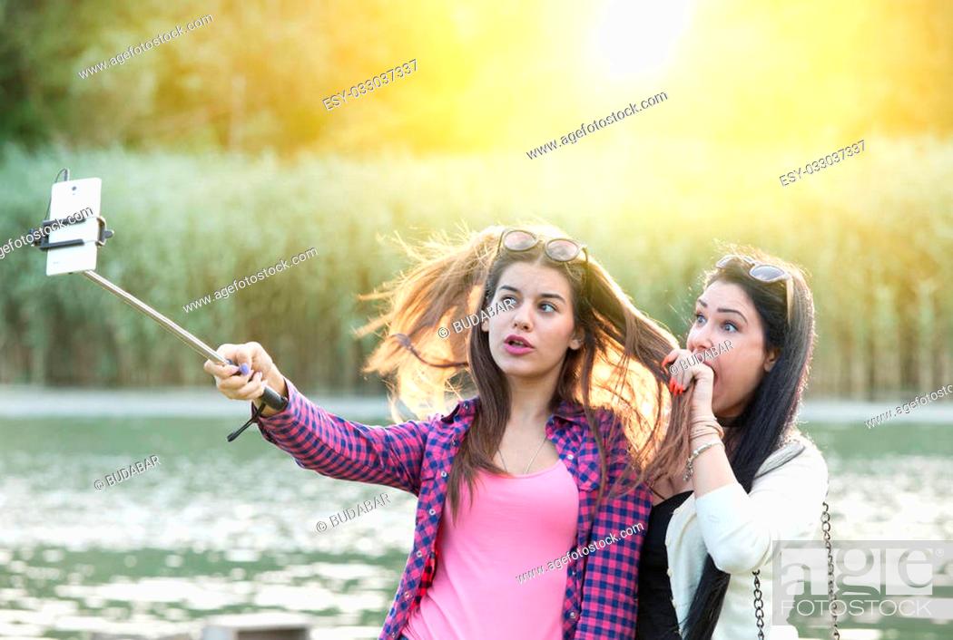 Stock Photo: Two pretty teenage girls taking selfie on mobile phone with stick in the nature beside river. Positive and crazy emotions.