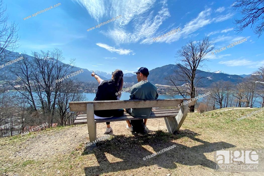 Stock Photo: Young couple takes a rest on a bench. Hikers on the Hoehenweg over the Tegernsee with a view of Rottach Egern on April 1st, 2021.