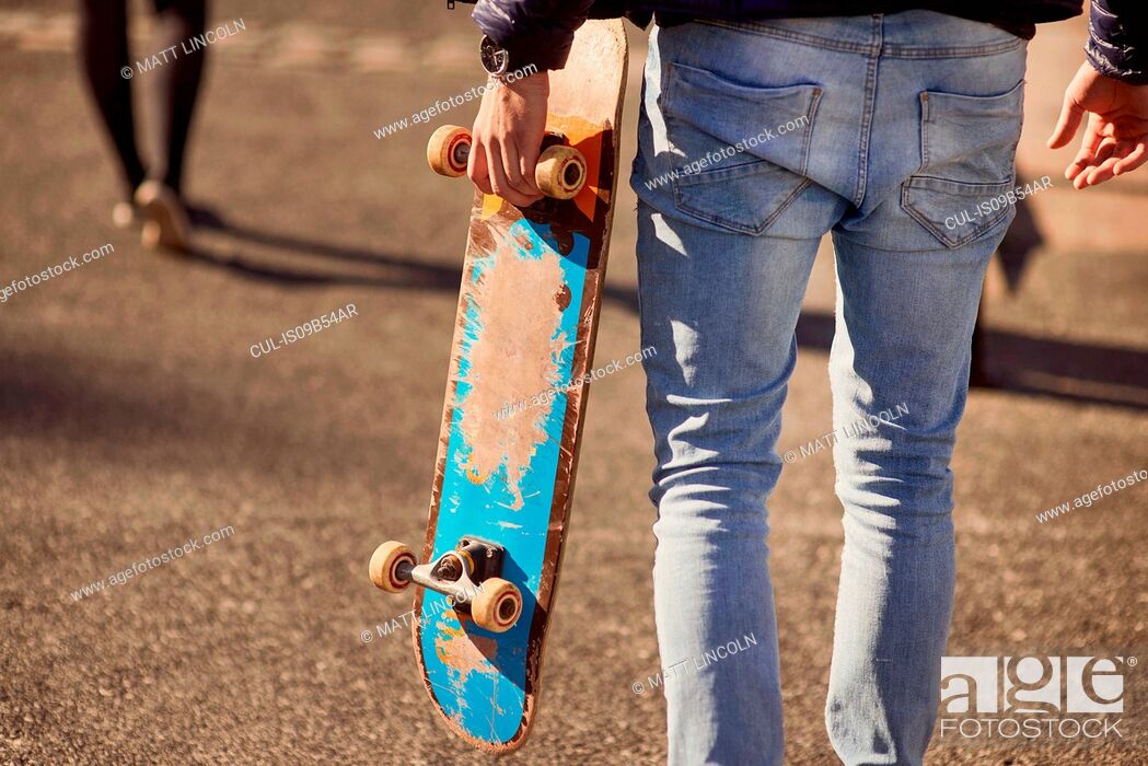 Stock Photo: Young man walking outdoors, carrying skateboard, rear view, low section, Bristol, UK.
