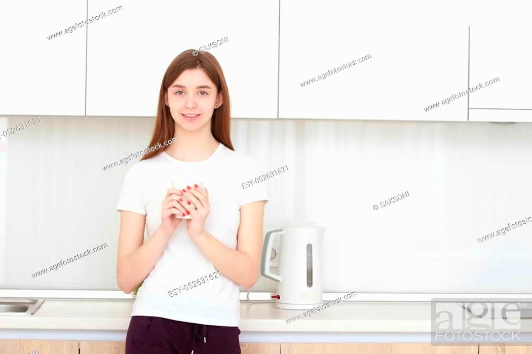Stock Photo: Pretty young woman with cup of coffee standing in kitchen and smiling. Real interior. Wide angle.
