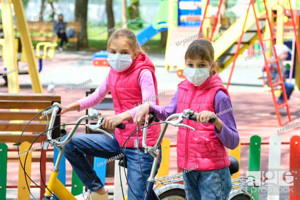 Stock Photo: Two girls in medical masks ride bicycles in the playground.