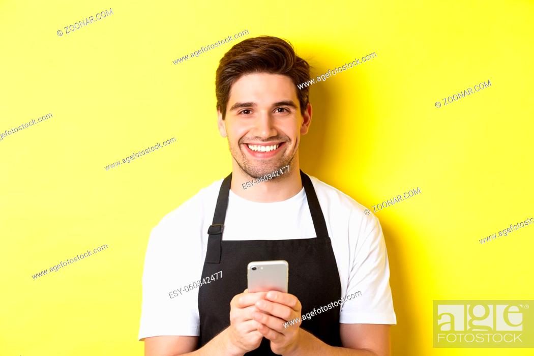 Stock Photo: Close-up of handsome barista sending message on mobile phone, smiling happy, standing over yellow background.