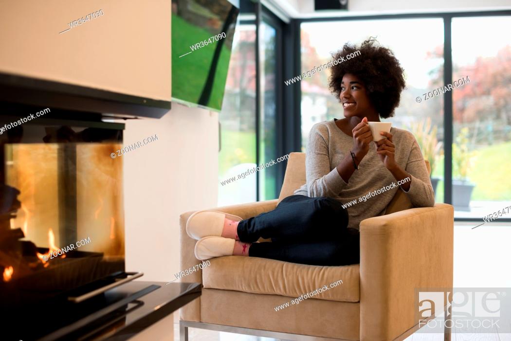 Stock Photo: Happy joyful african american woman drinking cup of coffee relaxing at fireplace. Young black girl with hot beverage heating warming up.