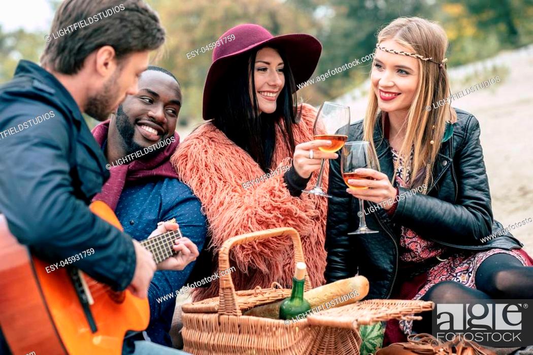 Stock Photo: Happy smiling friends holding wine glasses and enjoying guitar while sitting on sandy beach at alfresco picnic.