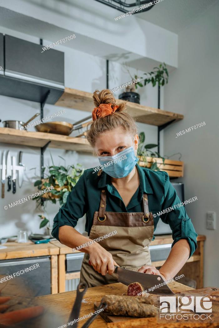 Stock Photo: Young female chef wearing protective face mask cutting salami while working in kitchen during COVID-19.