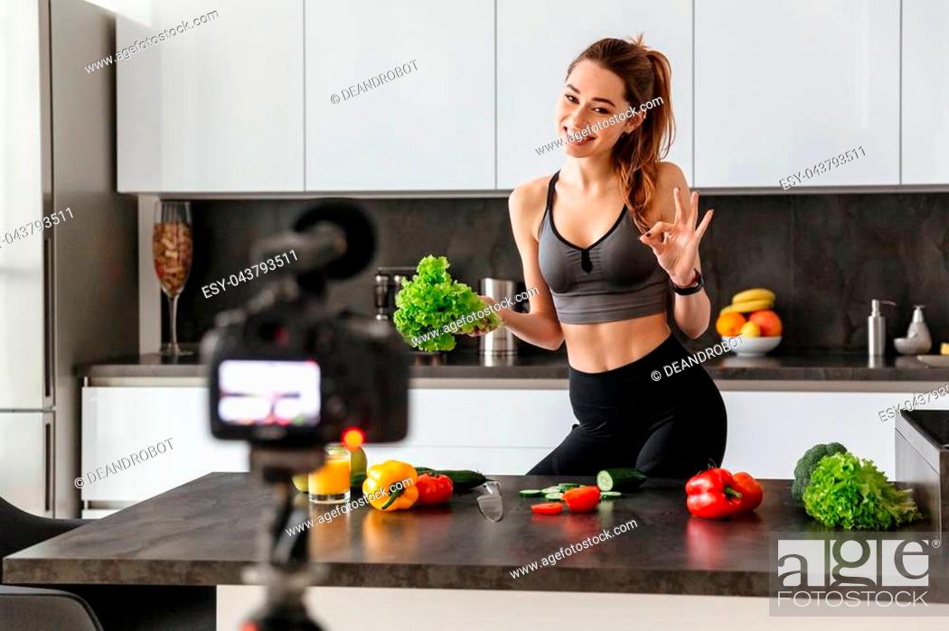 Photo de stock: Happy healthy young girl recording her blog episode about healthy food diet while standing at the kitchen at home and showing ok gesture.