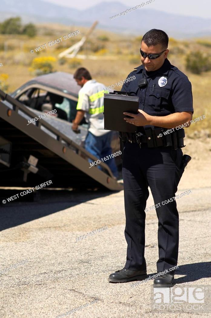 Stock Photo: Police officer writing notes tow truck driver lifting crashed car in background.