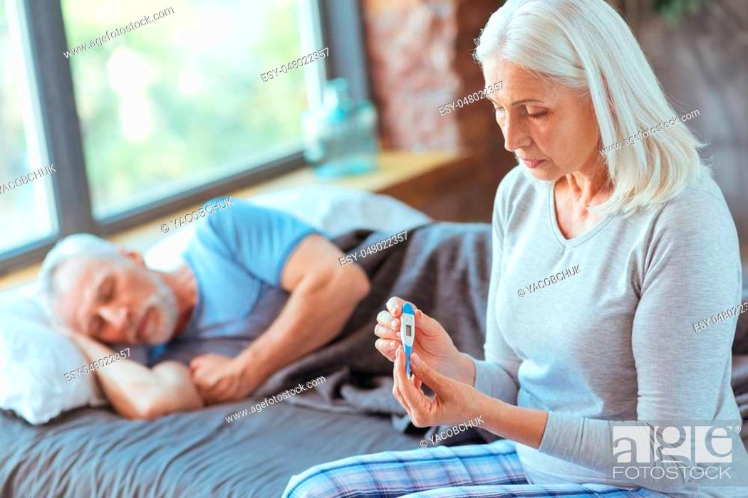 Stock Photo: Concerned about health. Sad aged woman holding a thermometer and sitting on bed while her sick husband sleeping in the background.