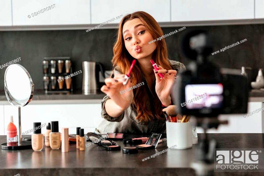 Stock Photo: Attractive healthy young girl recording her video blog episode about new cosmetic products while sitting at the kitchen table at home and applying make-up.