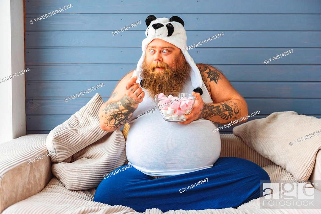 Hungry fat man is looking at zephyr with appetite. He is relaxing on sofa  at home in funny panda hat, Stock Photo, Picture And Low Budget Royalty  Free Image. Pic. ESY-048149547 |